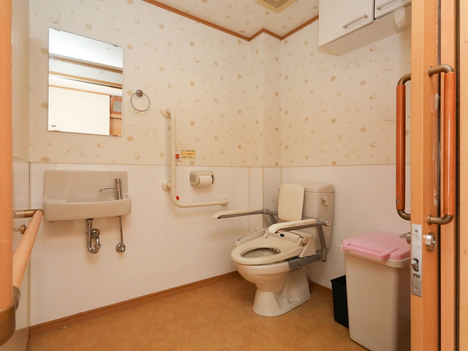 柄つきの壁紙が貼られた、洋式トイレである。手洗い場が備えられている。上部には鏡がある。