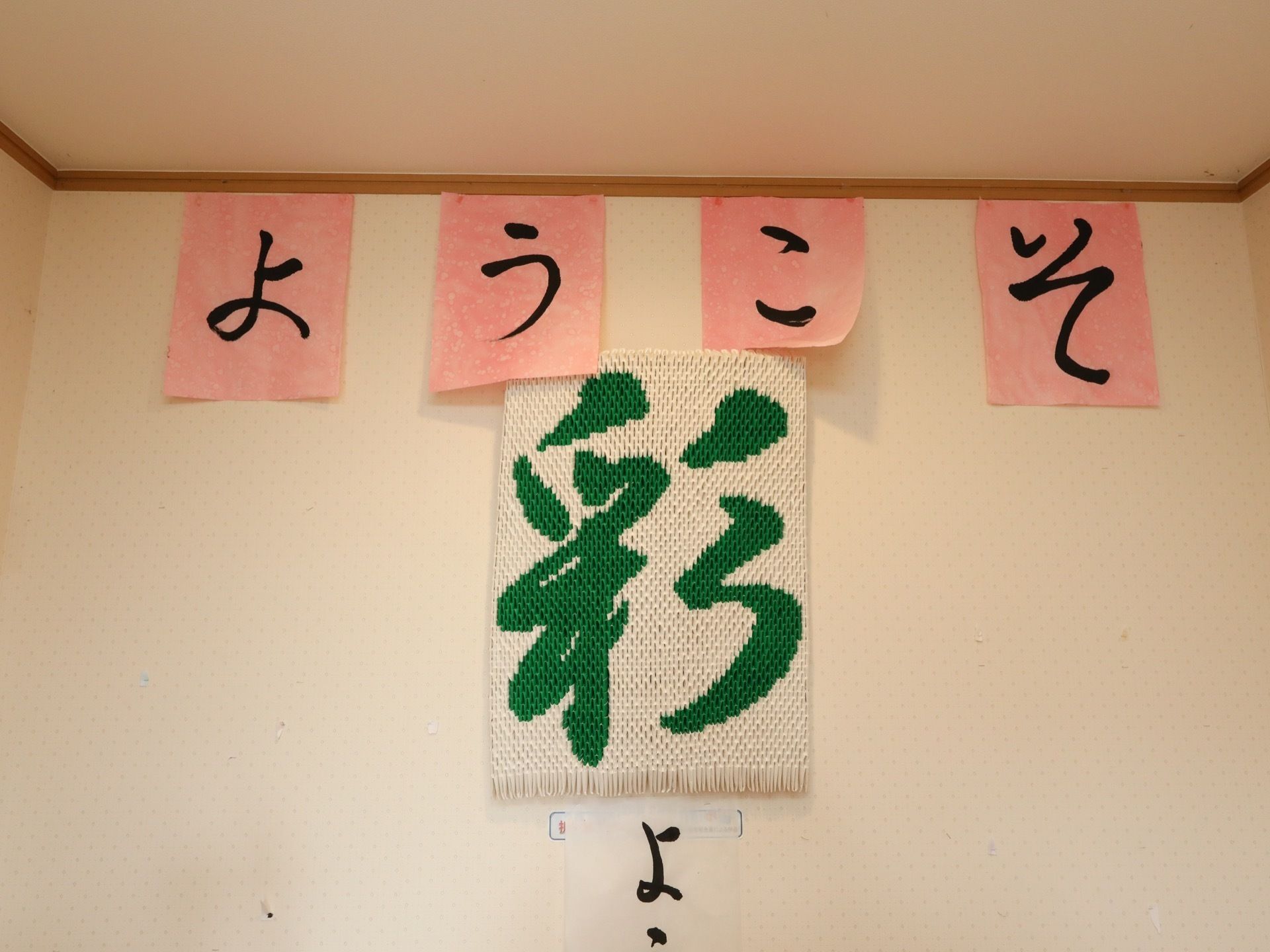 白い壁紙が貼られた、シンプルな一室。手書きで文字が書かれた紙が飾られている。真下には施設名がグリーンで大きく描かれている。