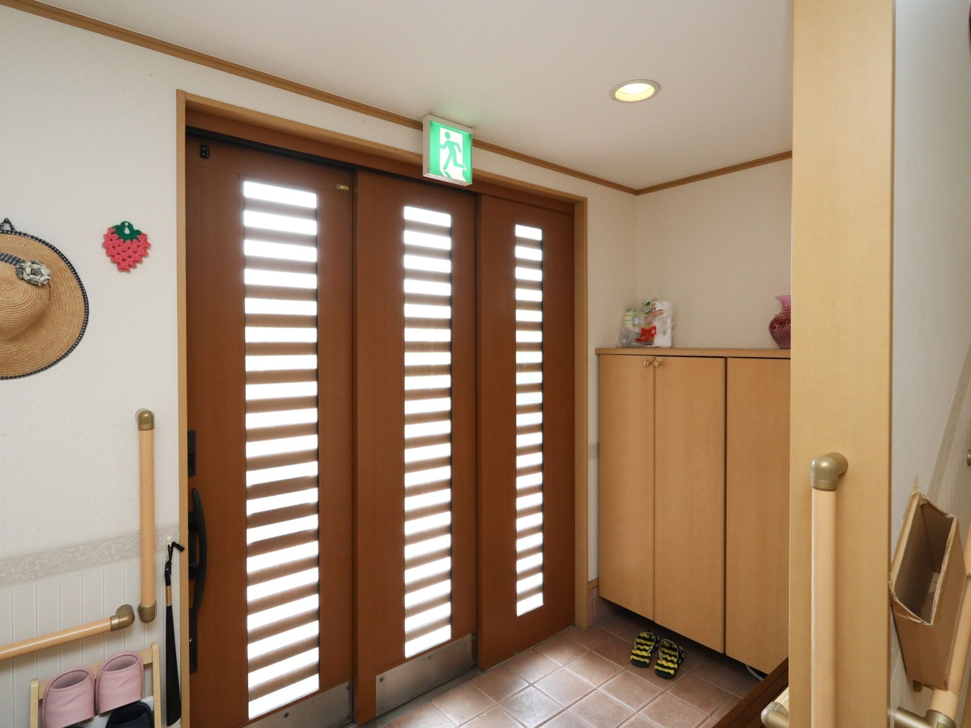格子のついた、３枚連動式の玄関扉がある。非常口誘導灯が上部にある。出入口に、手すりも備えられている。