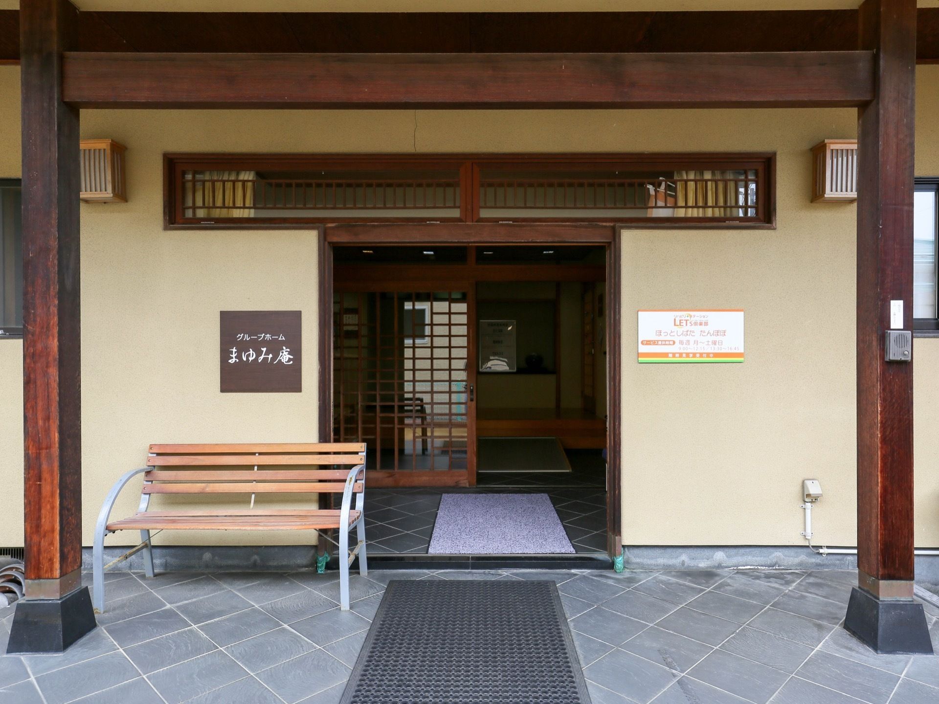 ２本の木の柱の間に施設の入口がある。入口の手前には滑り止めのマットが敷かれている。ドアの隣にはベンチがある。