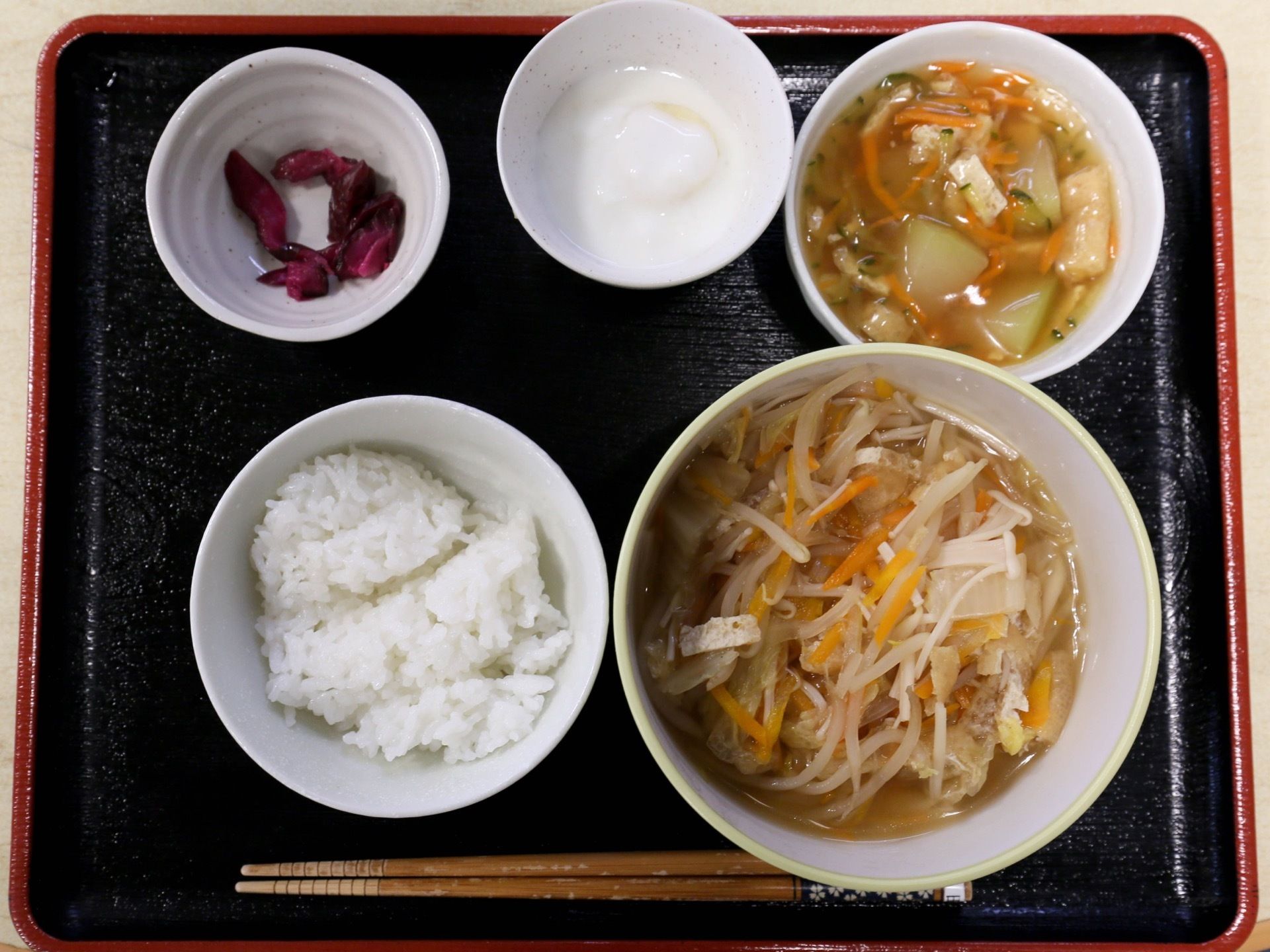 黒いお盆の上にお箸、ご飯、野菜の煮物、漬物、ヨーグルトなどが置かれている。それぞれ違う器に配膳されている。