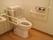 白色の便座を完備したトイレである。Ｌ字型や可動式の手すりを完備している。背中がもたれられるクッションもある。