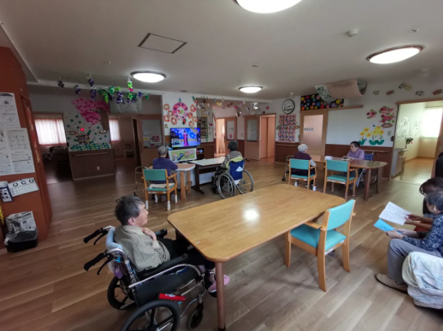 なにかを見て笑顔で手をたたいている高齢女性とスタッフの様子。施設での一場面の写真