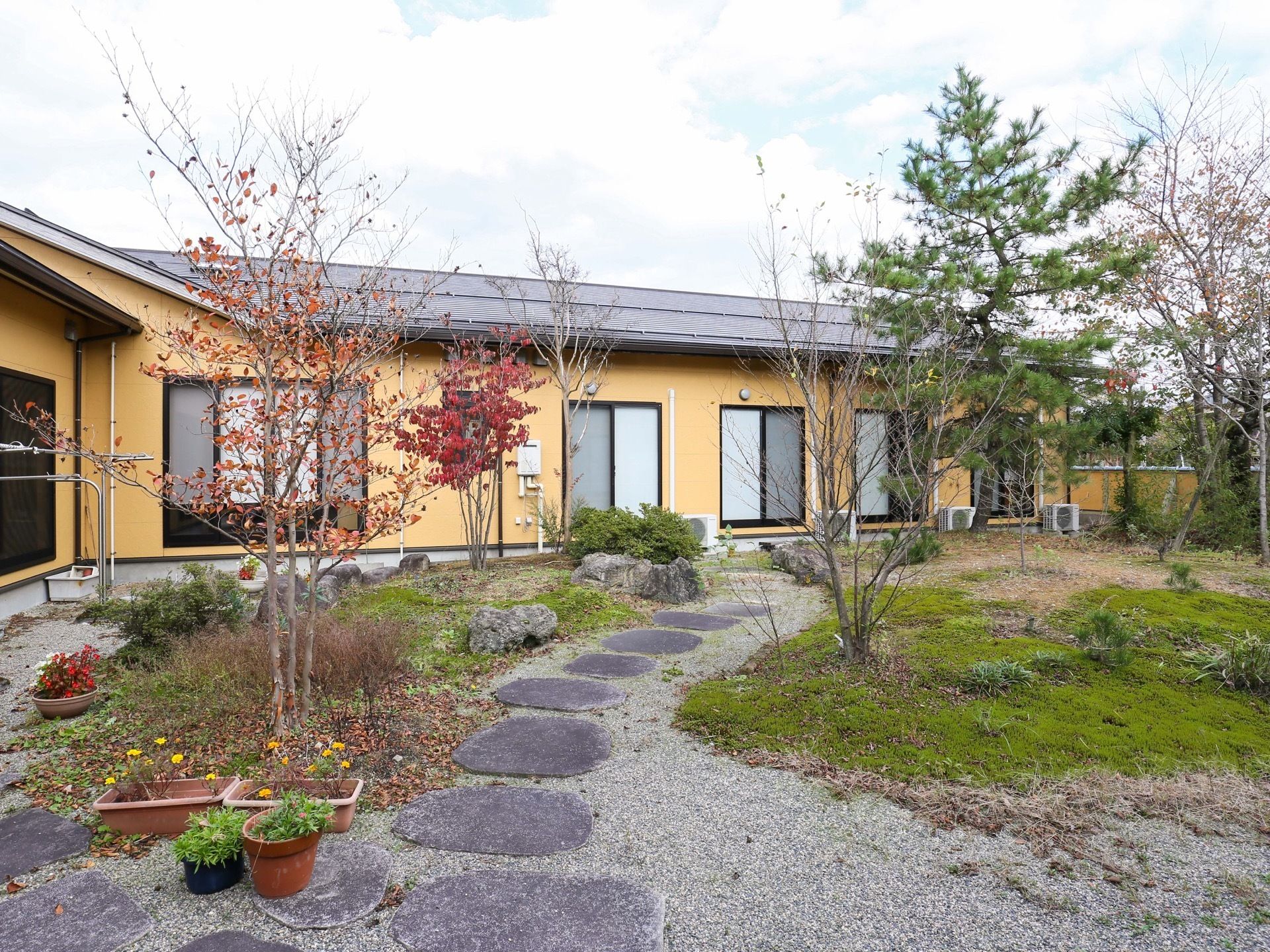 オレンジの外壁にグレーの三角屋根が乗った平屋づくりの建物である。建物の前は庭が広がり、飛び石や樹木がある。