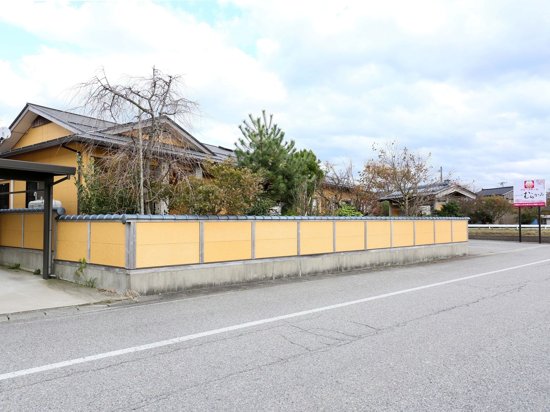 建物の外壁と同じ色のフェンスで囲まれており、その内側に施設が見えている。建物のフェンスの間にたくさんの樹木が植えられている。