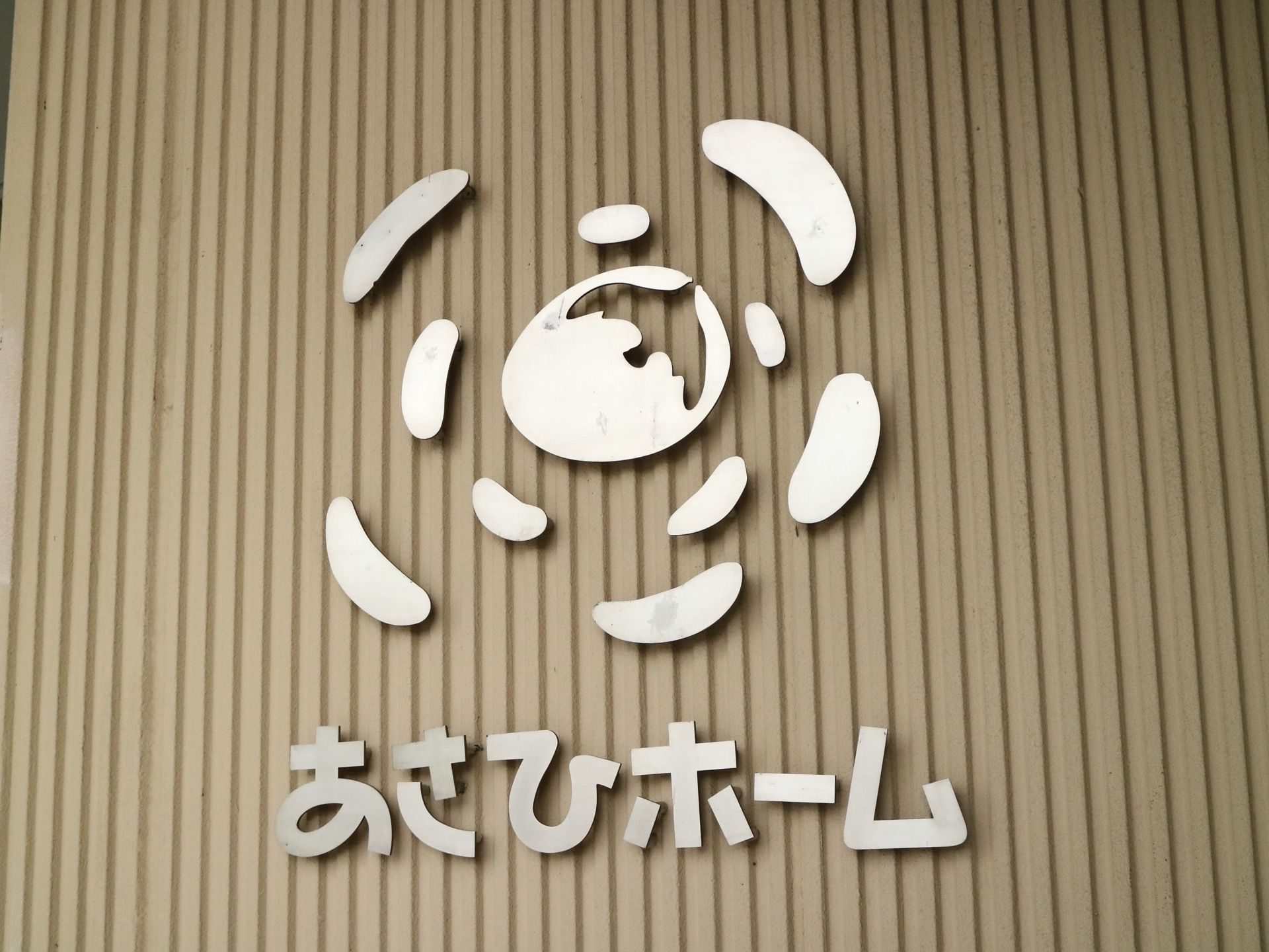 薄い茶色の縦縞模様の外壁に取り付けられた施設名の看板である。ステンレス製で、施設名の他、ロゴも入れられている。
