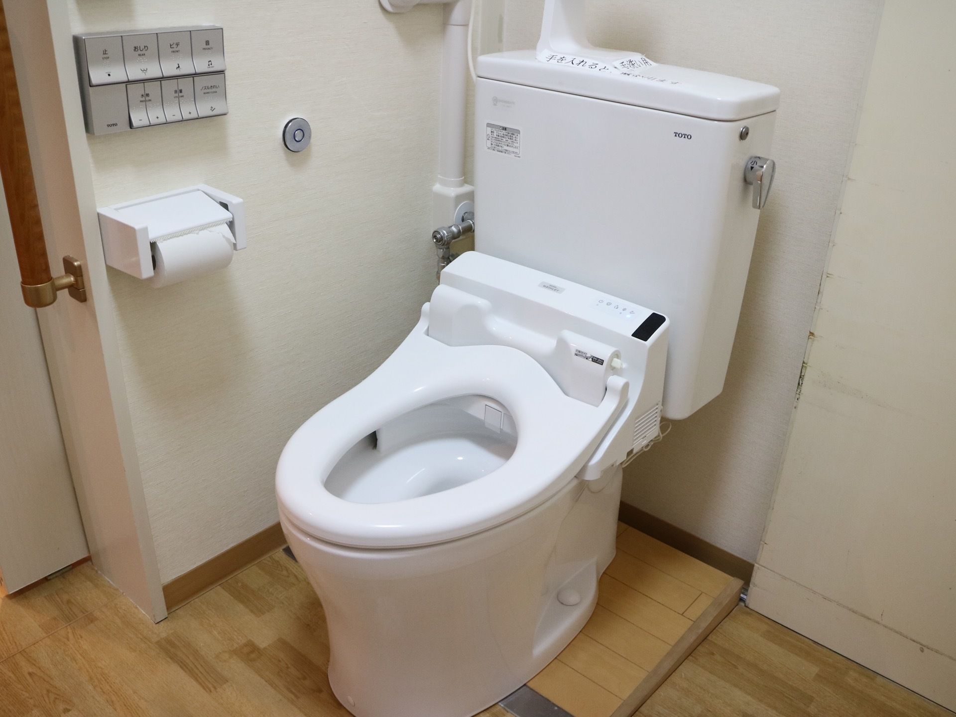 白色の洋式便座が備え付けられた、トイレである。壁にリモコンやトイレットペーパーホルダーが設置されている。