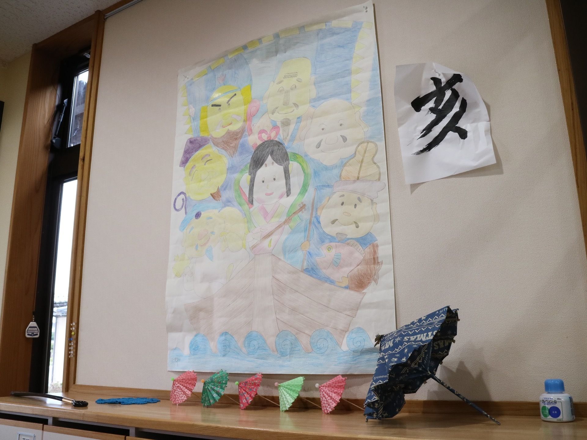 壁に貼られた入居者様の作品である。宝船の七福神と習字作品で、その前にはミニチュアの傘が並び、見る人の心を和ませる。