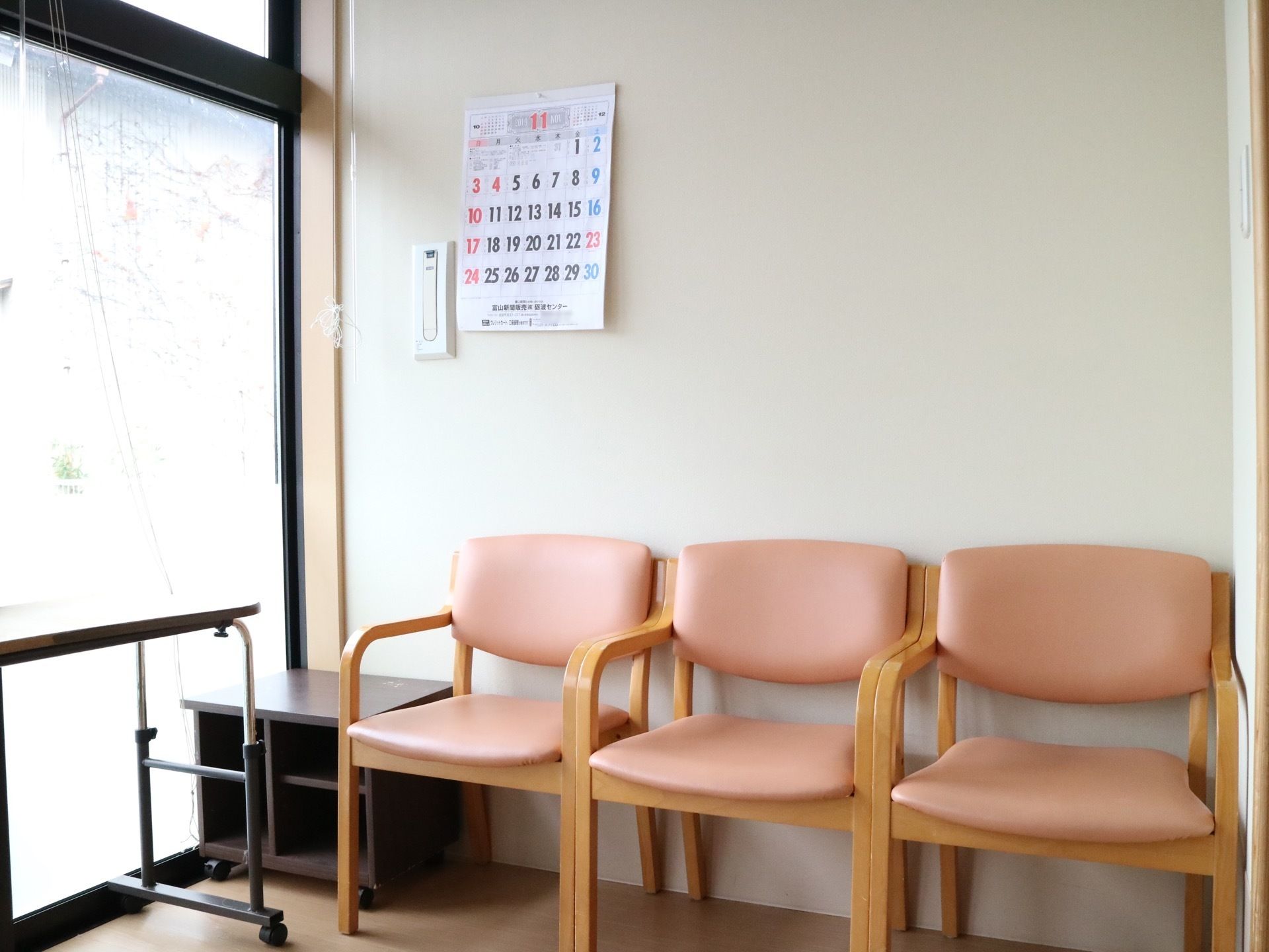 廊下の突き当りで、左手は窓があり外光が差し込んでいる。正面の壁際にピンク色の椅子が３脚並べられている。