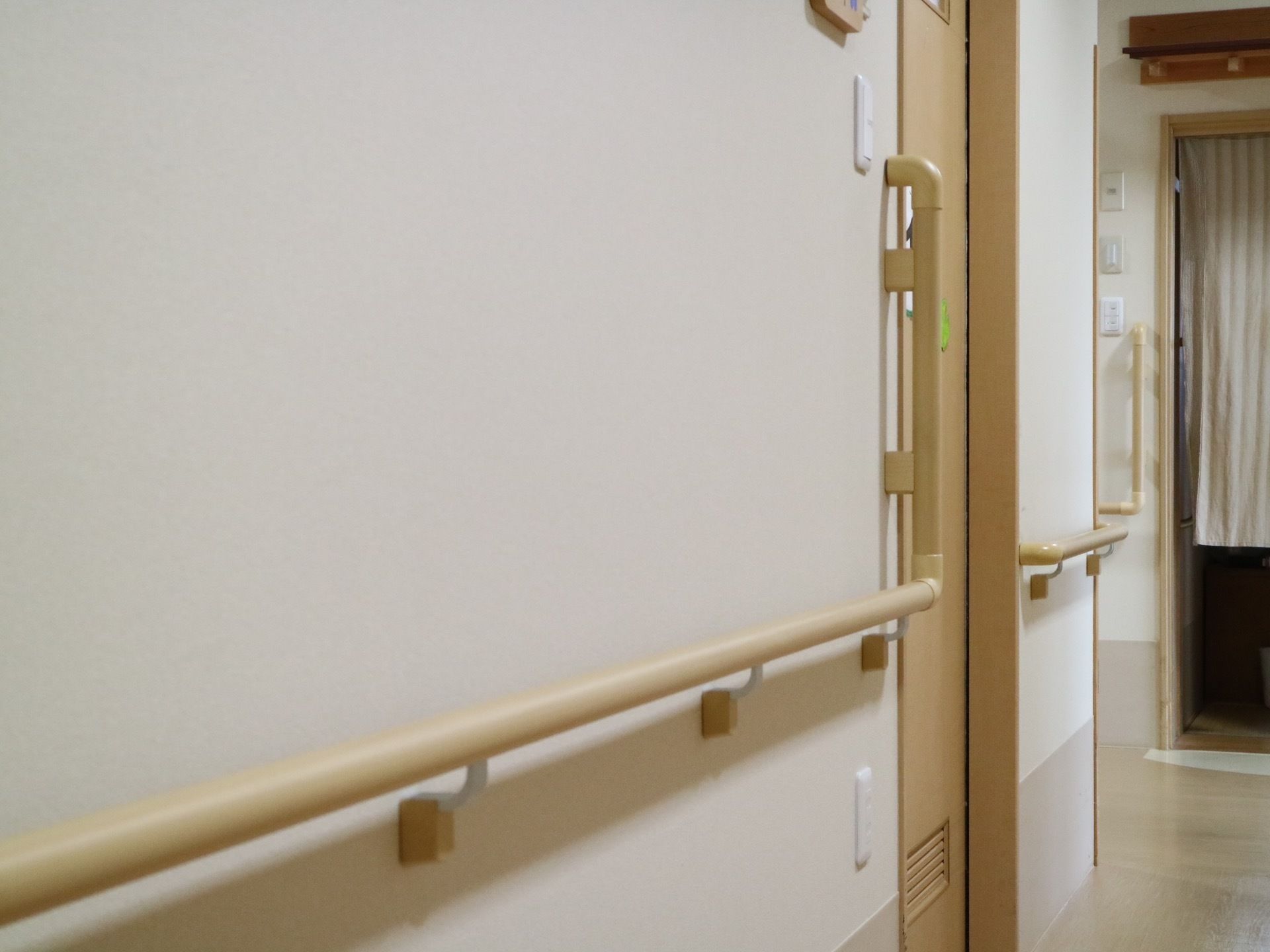 白い壁の廊下に設置された手すり。腰ほどの高さに木製の手すりが取り付けられている。右奥に向かって廊下が伸びている。