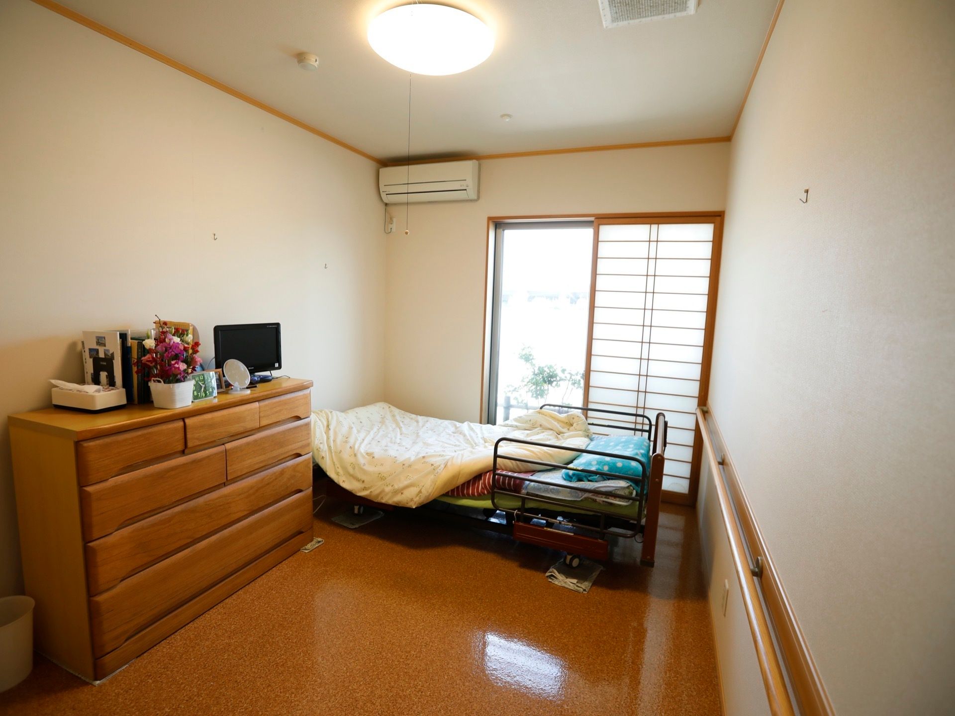 フローリング仕様の個室である。床色は落ち着いたブラウン調。同系色のチェストがある。ベッドもある。