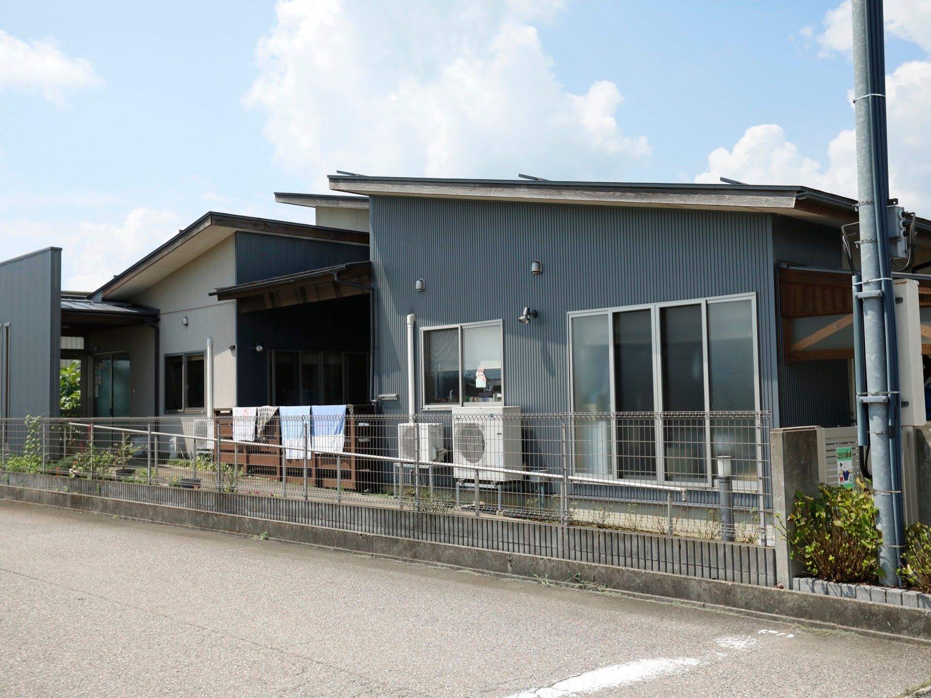 グレーを基調とした、シックな外観。そばに電柱が立っている。敷地はフェンスで囲まれていて、目の前は道路になっている。