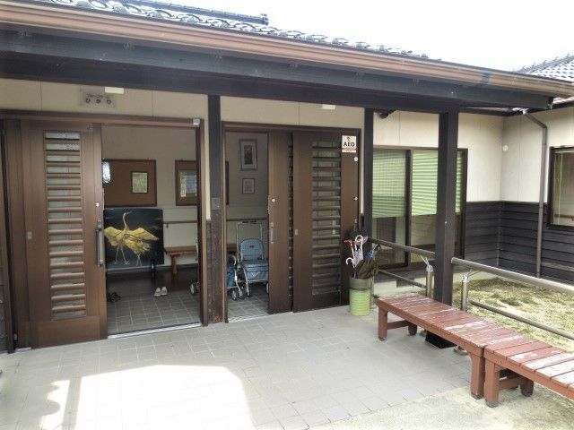 平屋建ての建物の玄関に木製のベンチが2台置かれている。入り口には傘立てと鶴が描かれた置物が置かれている。