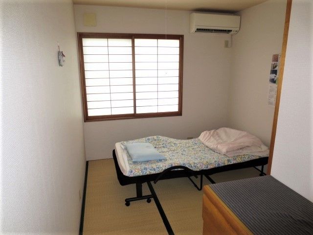 畳張りの居室に障子の貼られた窓がある。ベッドには水色の枕とピンク色の掛け布団が置かれている。