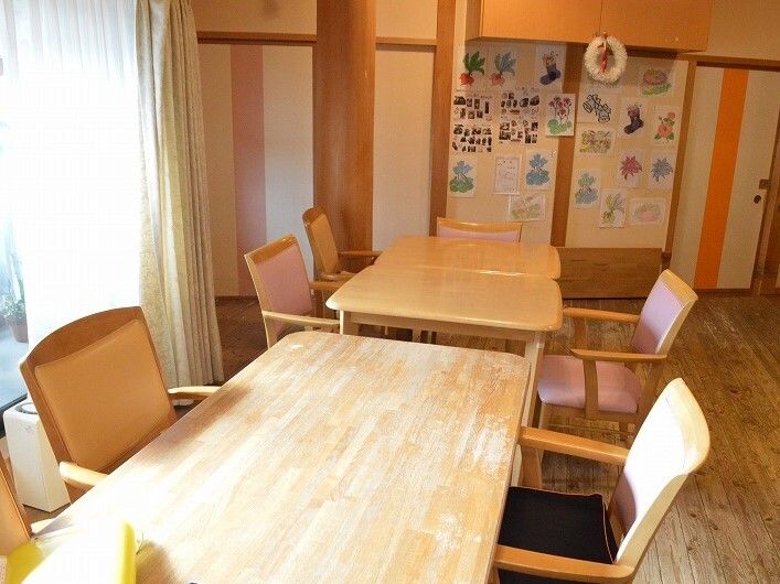 木製のダイニングテーブルセットが複数置かれたリビングダイニング。壁には入居者の作品と思われる塗り絵が飾られている。