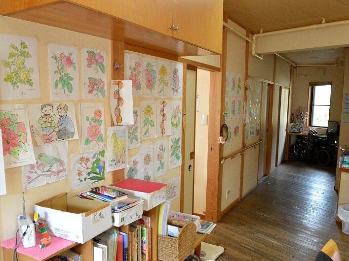 入居者が塗ったと思われる塗り絵がたくさん張られた廊下の壁。廊下はまっすぐで、本棚の上にはPHSも置かれている。