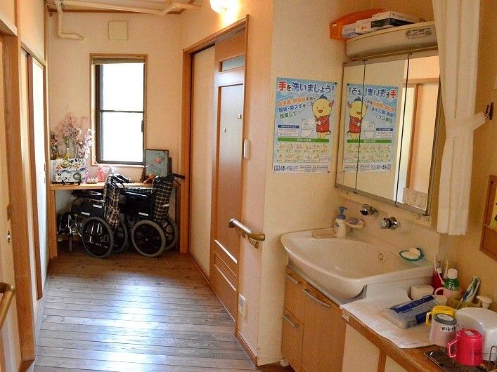 三面鏡タイプの収納が付いた大きな洗面所があり、横には歯ブラシが置かれている。廊下の突き当りには窓があり、車いすも置かれている。