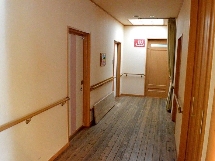 白い壁には手すりが付き、フロアはフローリングで、両サイドに居室のドアがあり、突き当りのドアの横には温泉マークが付いている。