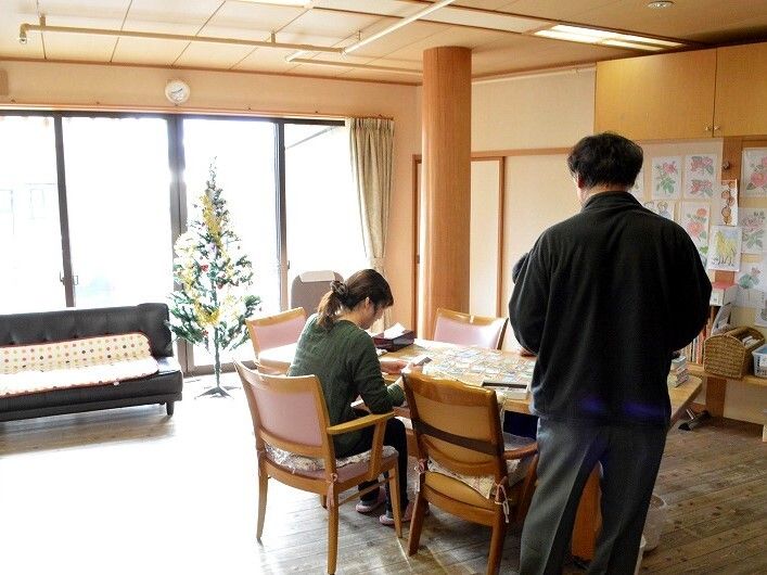 カルタで遊んでいるスタッフと入居者。男性が一人テーブルのそばに立っている。窓のそばにはクリスマスツリーがある。