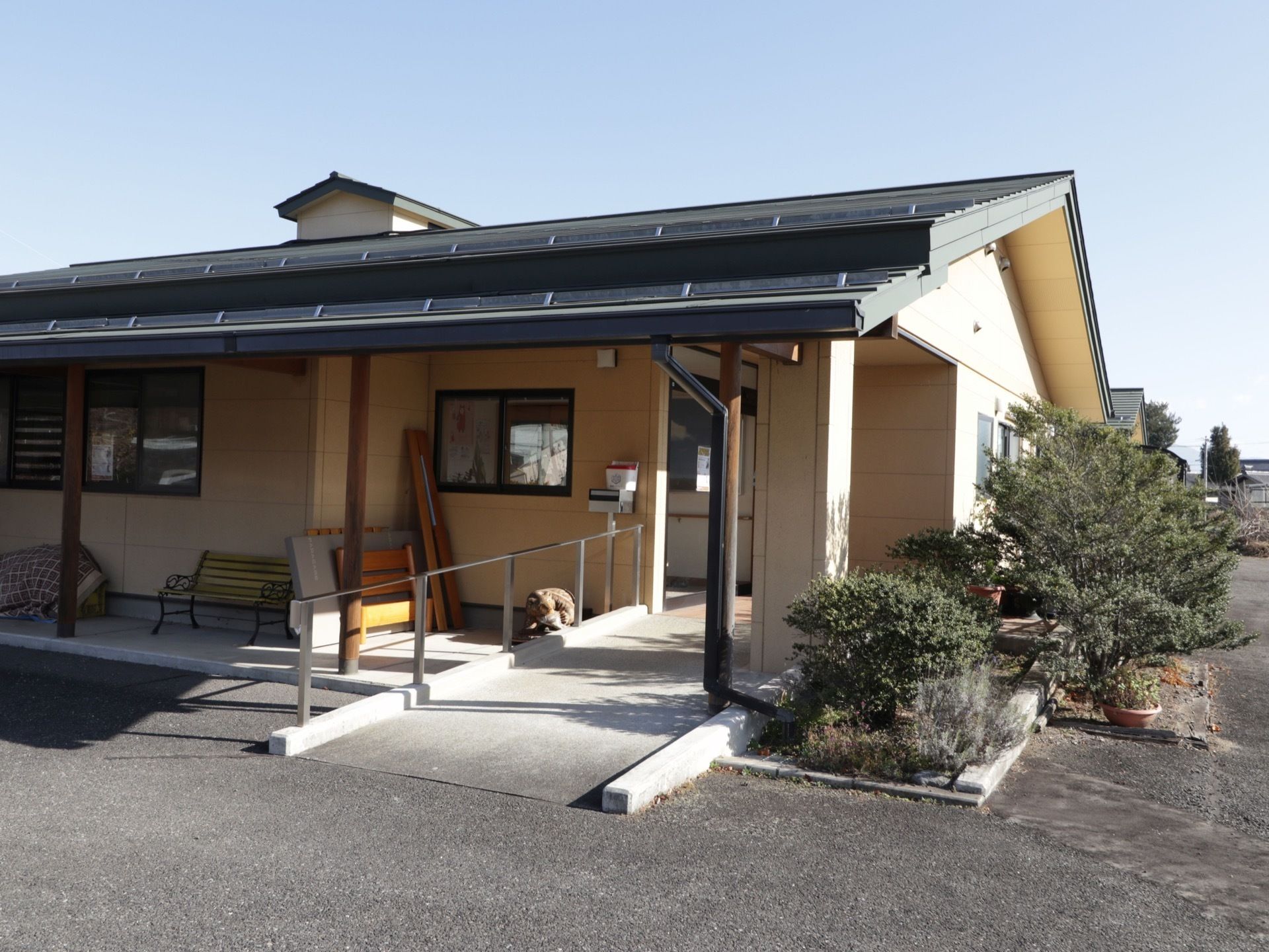 施設は和風の平屋づくりになっている。玄関横の壁のそばにベンチが置かれている。背丈の低い木が植えられている。