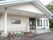 三角屋根の付いた白い建物がある。玄関に向かって手すりの付いたスロープが付いている。地面には植物が植えられているプランターが並んでいる。