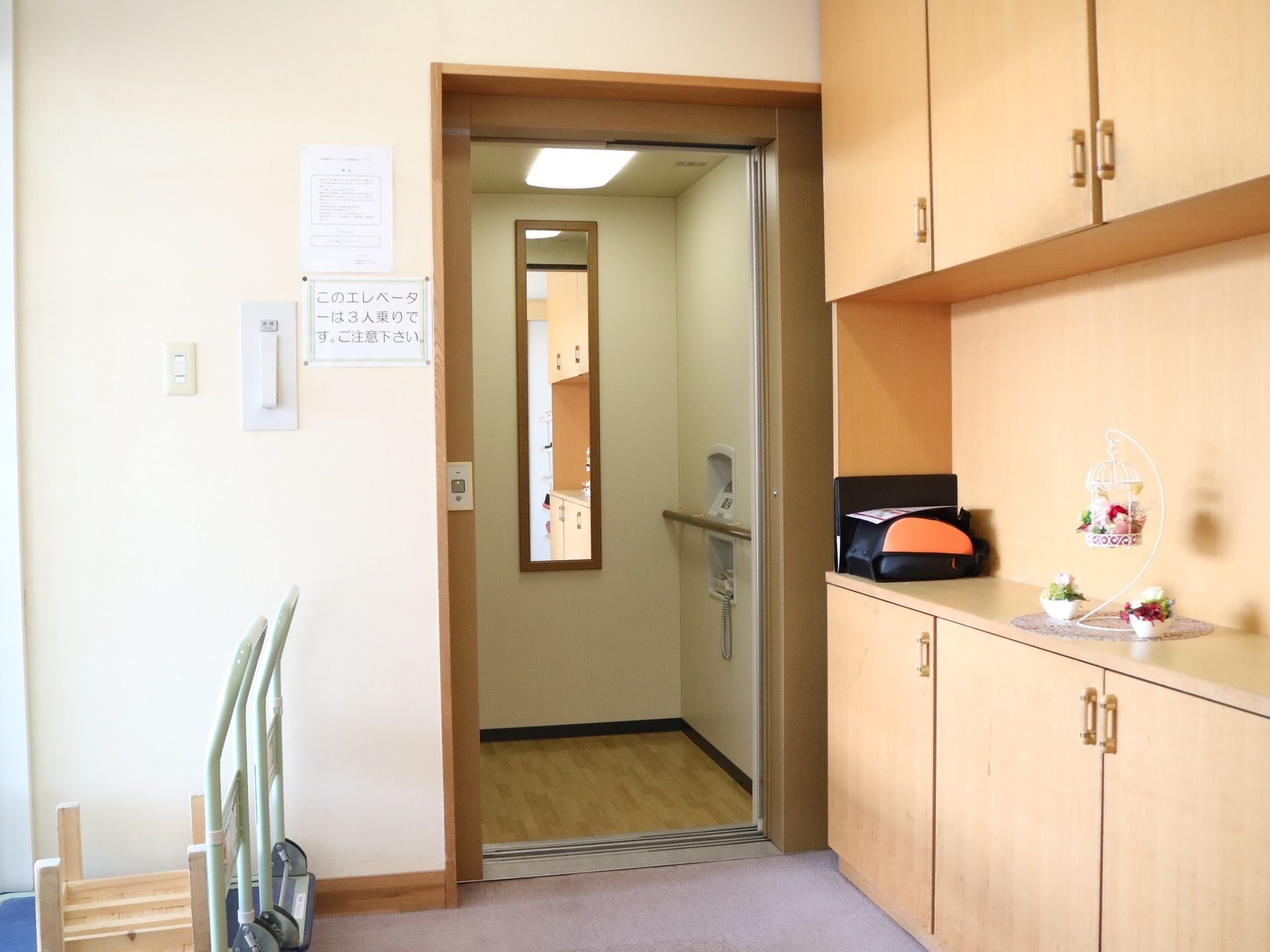 右手の壁に天井まで届く収納スペースがあり、オブジェが飾られている。正面にはエレベーターがあり扉が開いている。