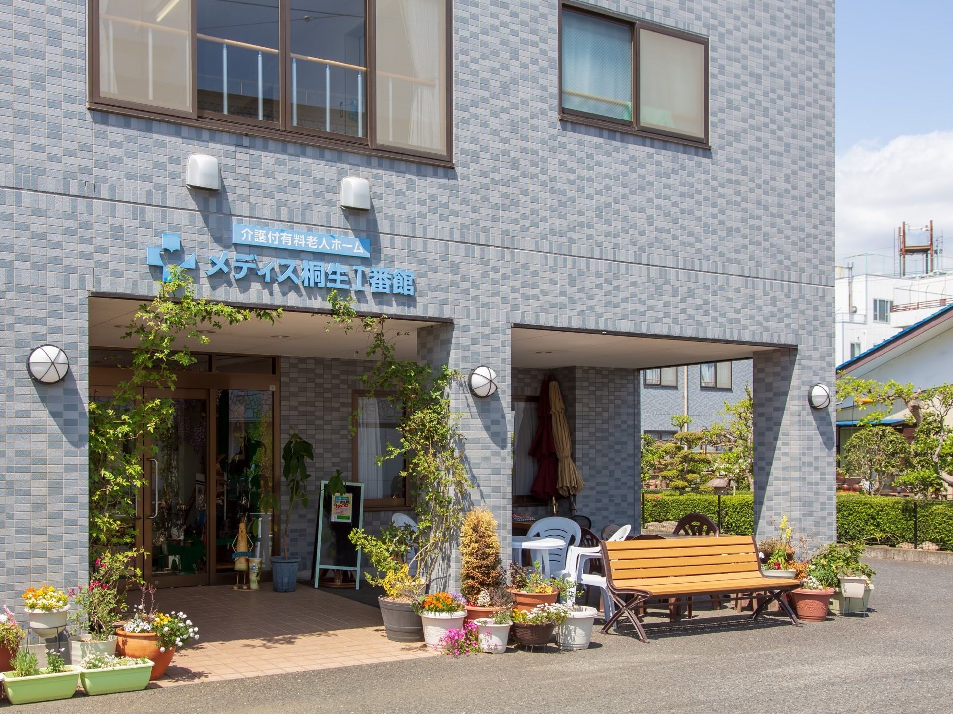 ブルー系の煉瓦調の外壁でおしゃれな建物である。玄関前には植物が置いてあり、ベンチを設置しているので、ひなたぼっこができる。