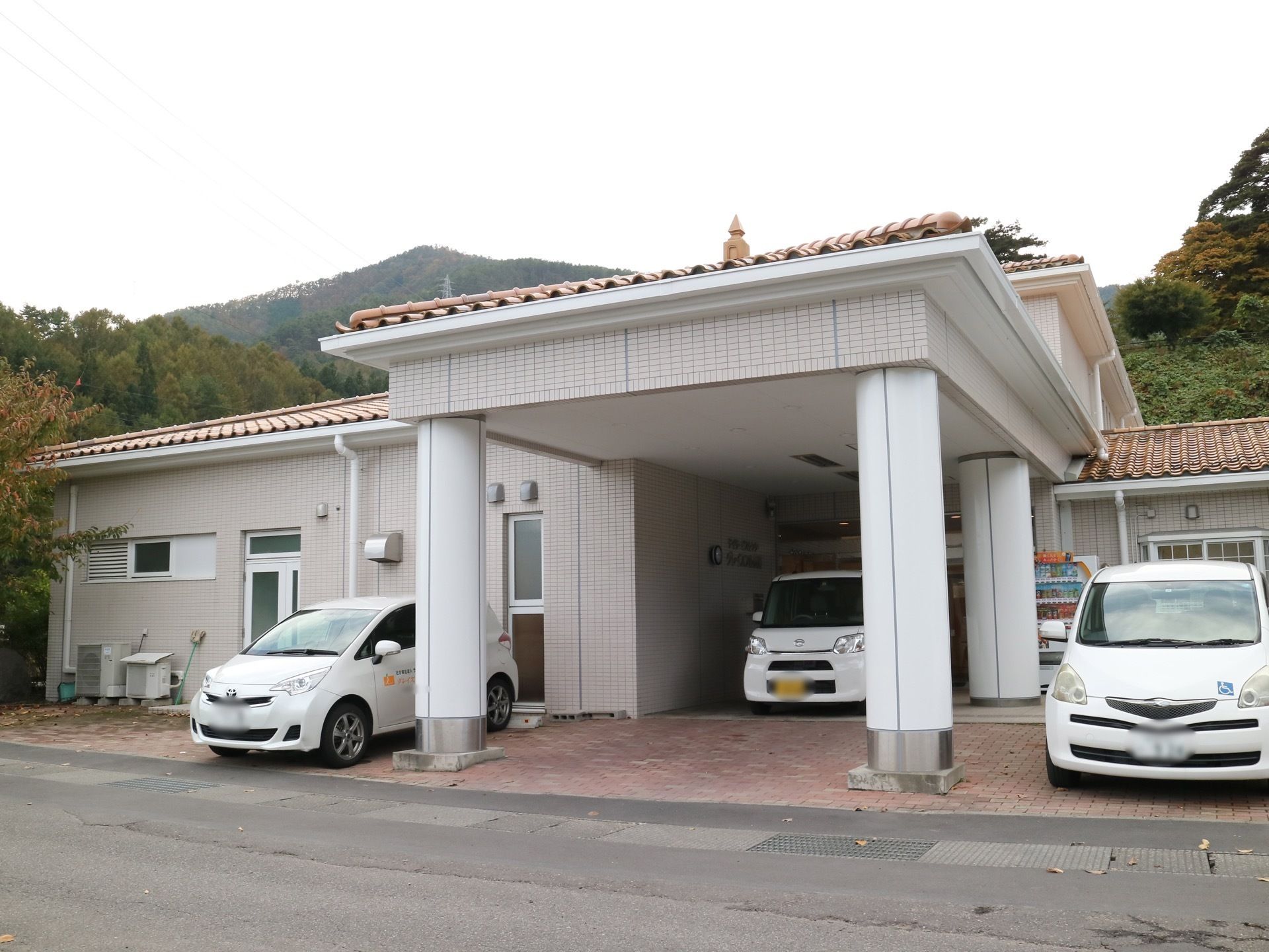 白い大きな柱が２本立っており、玄関ポーチの屋根を支えている。屋根の奥には軽自動車が停まっており、柱の横にも車がある。