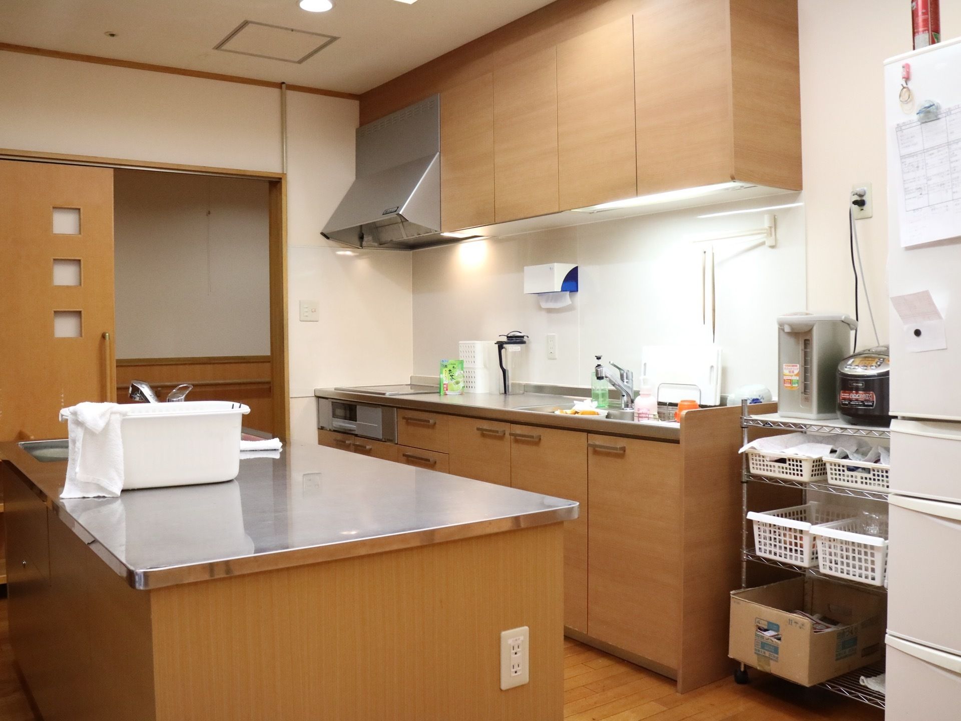 木目調の温かみのあるキッチンである。中央に独立したシンクがあり、電磁調理器仕様である。そばにはポットや炊飯器、冷蔵庫などが並んでいる。