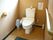 温水洗浄便座が設置されたトイレが中央にある。床には掃除道具やごみ箱が置かれている。背後の壁に貼り紙がされている。