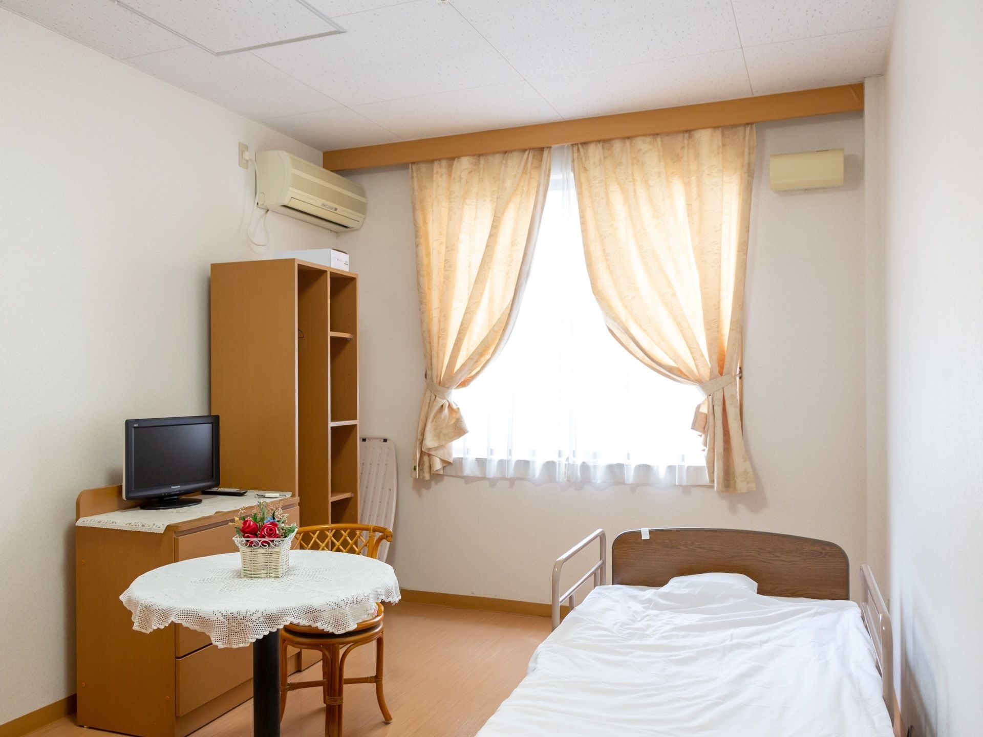 収納付きのテレビ台、本棚、丸テーブルとイス、介護ベッドが設置されている。居室にはエアコンが完備されている。