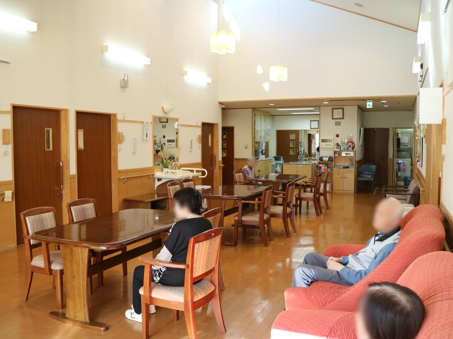 木製のテーブルセットや赤いソファがたくさん並べられた共有スペースで入居者様がのんびりとくつろいでいる。