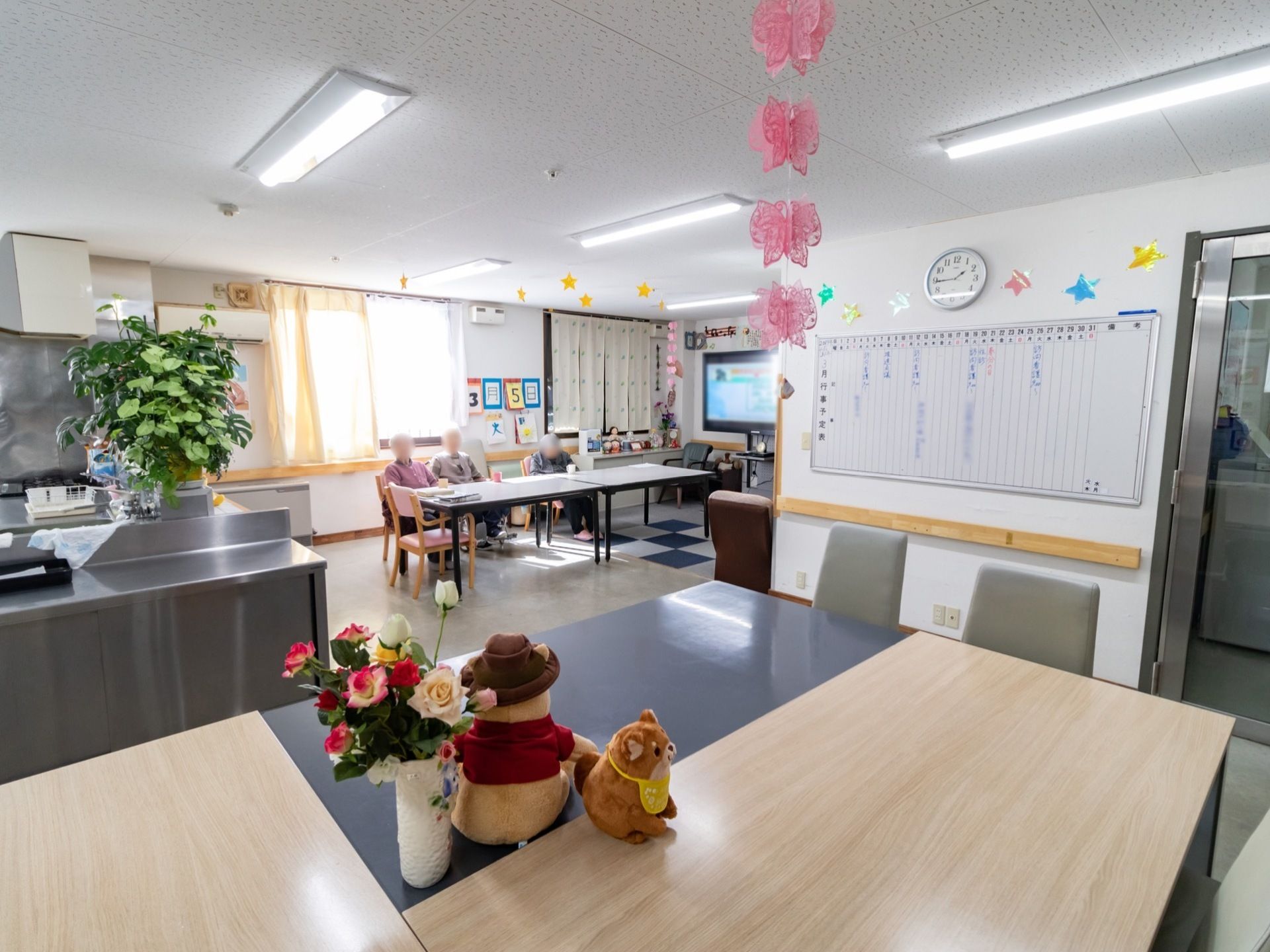 共有スペースでは入居者様がテレビを観ながら談笑している。手前のテーブルの上には花やぬいぐるみが飾れている。
