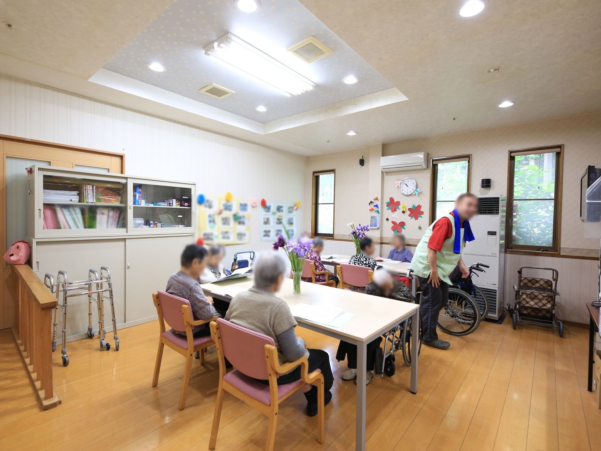 床がフローリングの部屋には、机と椅子が複数用意され、６、７人の入居者様が机に向かって座っている。壁面には、写真や手作りの紙細工が飾ってある。