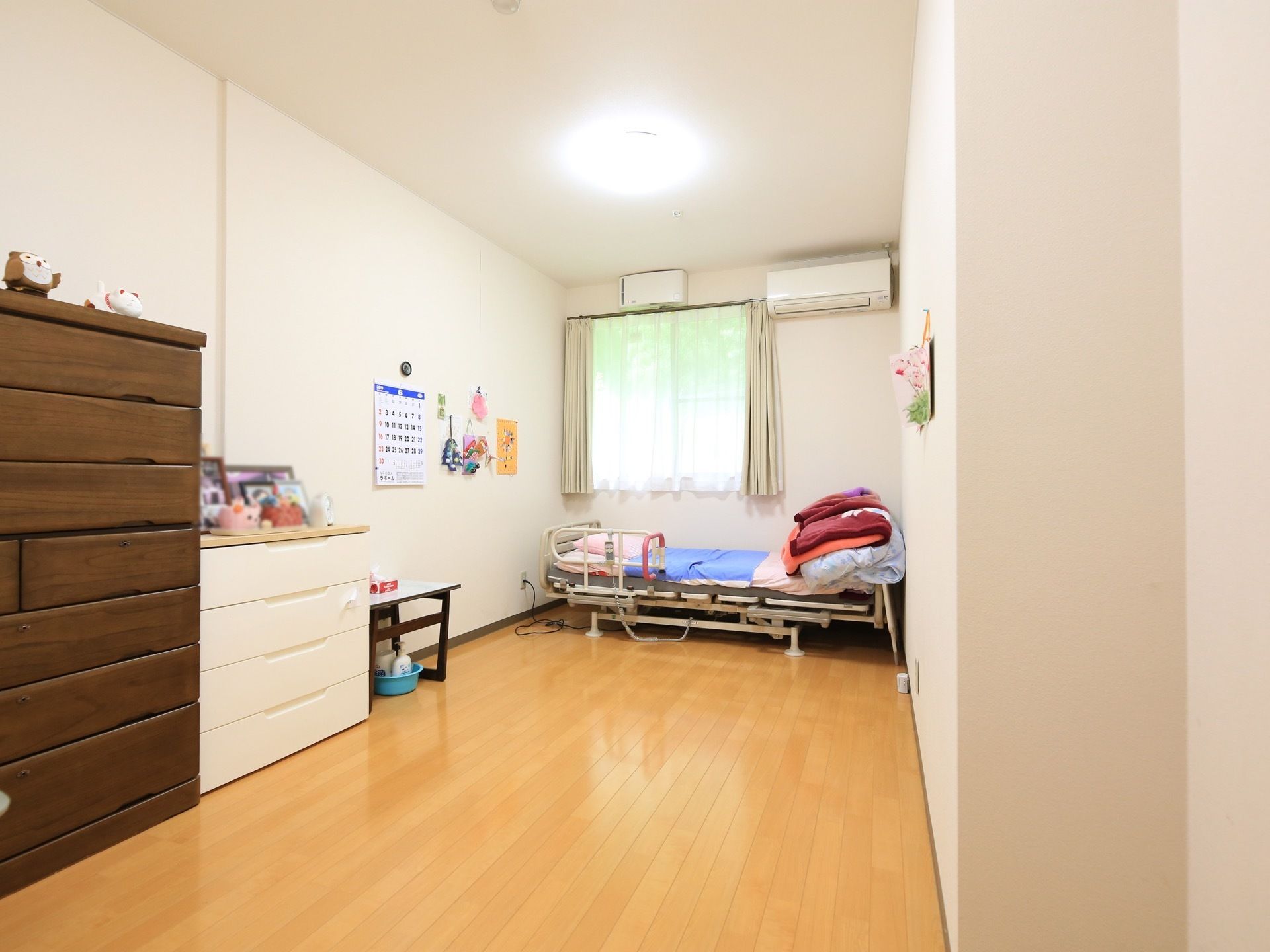 窓やエアコン、介護用ベッドが設置されている居室である。入居者様のお好みに家具が置かれ、壁にはカレンダーが貼られている。