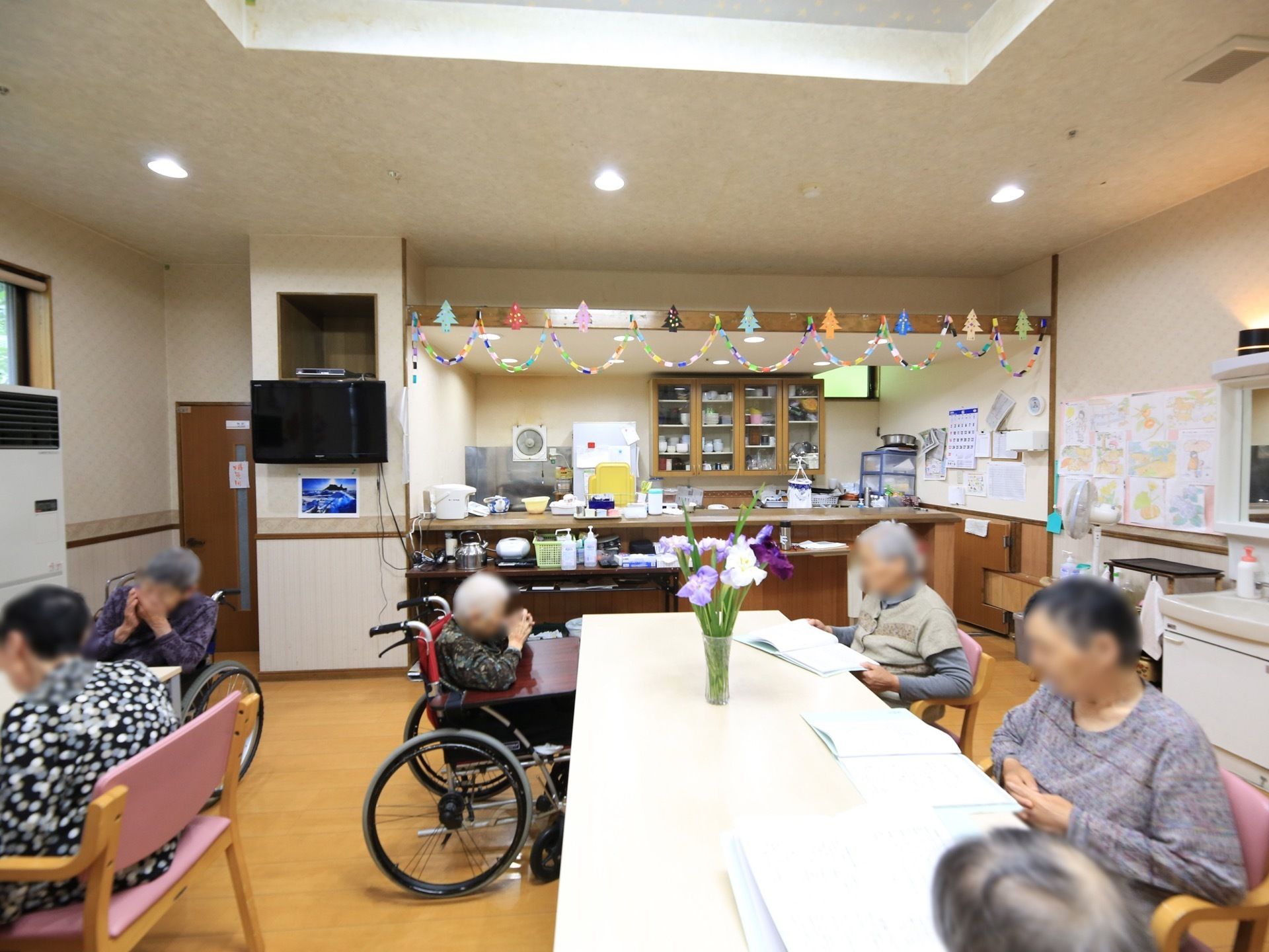 オープンキッチンや洗面台が完備されている食堂である。ダイニングテーブルの椅子に腰掛けている入居者様や車椅子をご利用の方の姿が見える。