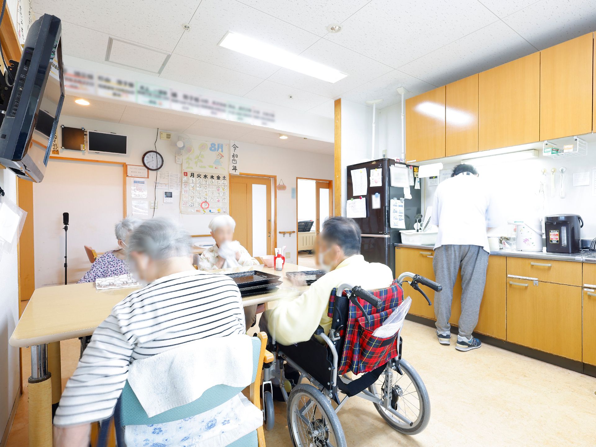 ブルーのエプロンを付けた若い女性スタッフに付き添われて原っぱのような場所を散歩している高齢女性。