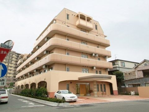 6階建ての大きな建物で、マンションのような雰囲気がある。入口の前には車を停めるスペースがあり、建物の横には植物が植えてある。