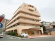6階建ての大きな建物で、マンションのような雰囲気がある。入口の前には車を停めるスペースがあり、建物の横には植物が植えてある。