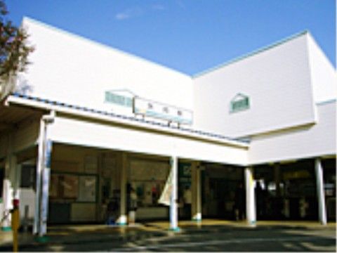 白い建物の駅があり、1階の屋根に看板が設置されている。中には券売機や改札口などがあり人が利用している。