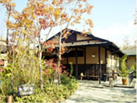 園路の途中に紅葉している樹があり、その前に平屋建ての日本家屋がある。切妻の屋根に壁は薄いベージュ色で腰高まで深い茶色の木が貼られている。