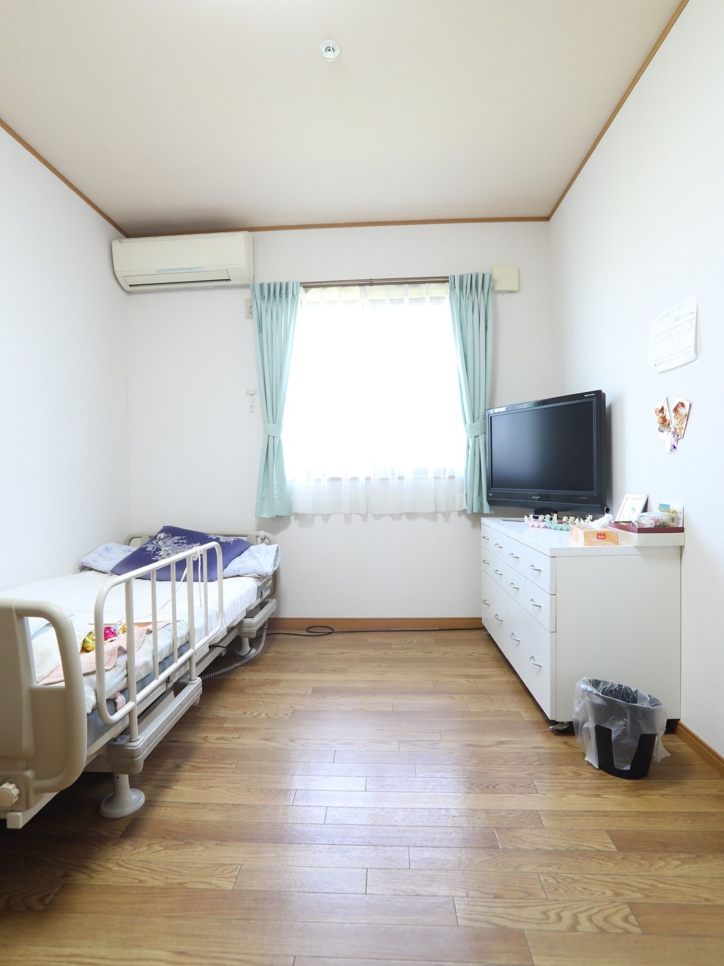 木目調のフローリングと白い壁の居室がある。カーテンの掛かった窓とエアコンがある。介護用ベッドと白い棚が置かれている。