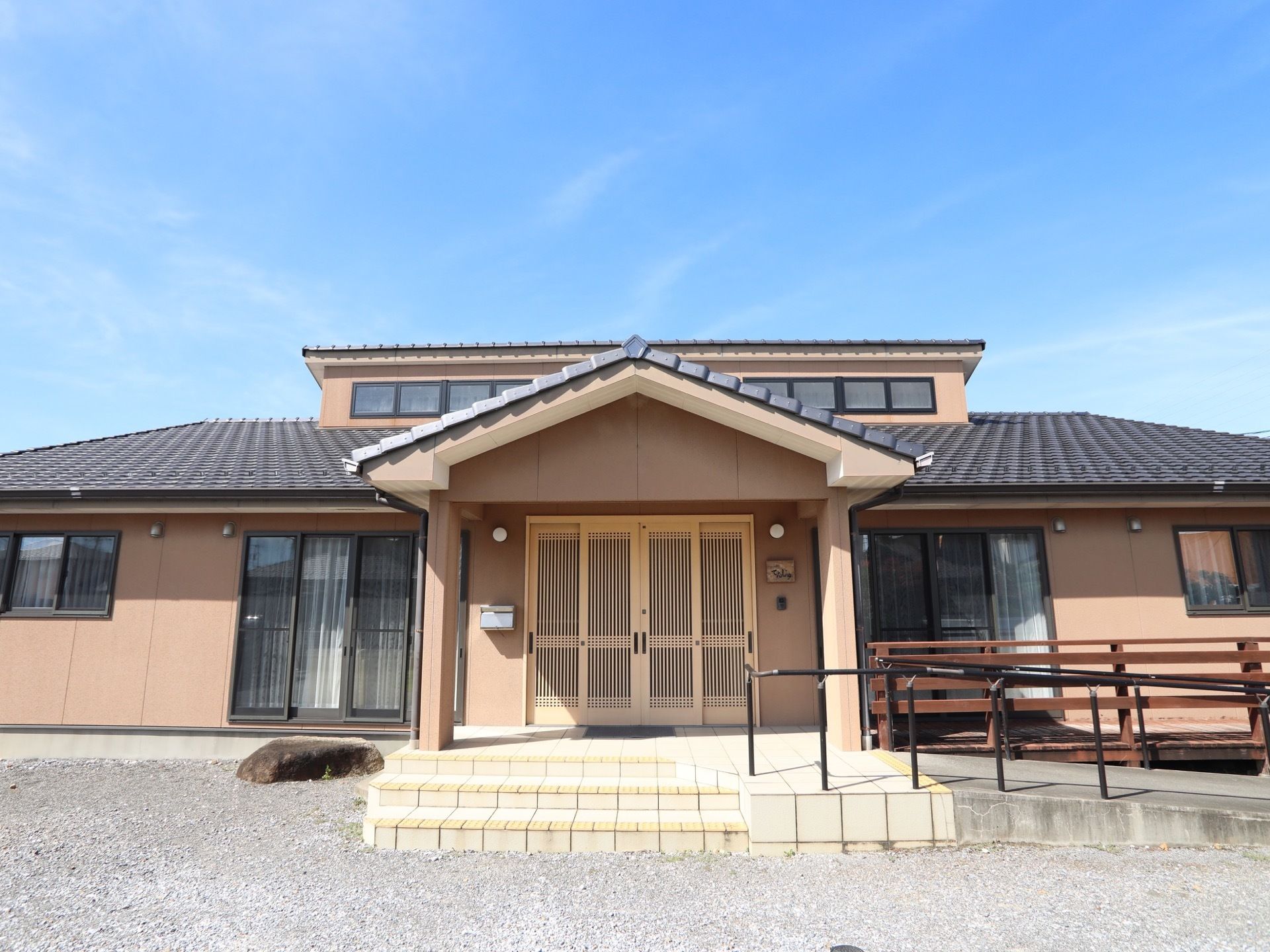 黒い瓦屋根のある、平屋建ての和風の建物がある。木の格子状の玄関があり、玄関に向かって手すりの付いたスロープがある。