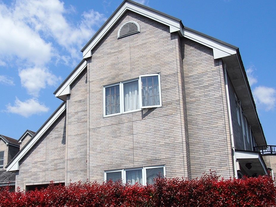 グレー系の外壁に三角屋根が付き、赤く色づいた生垣に囲まれた2階建ての建物。