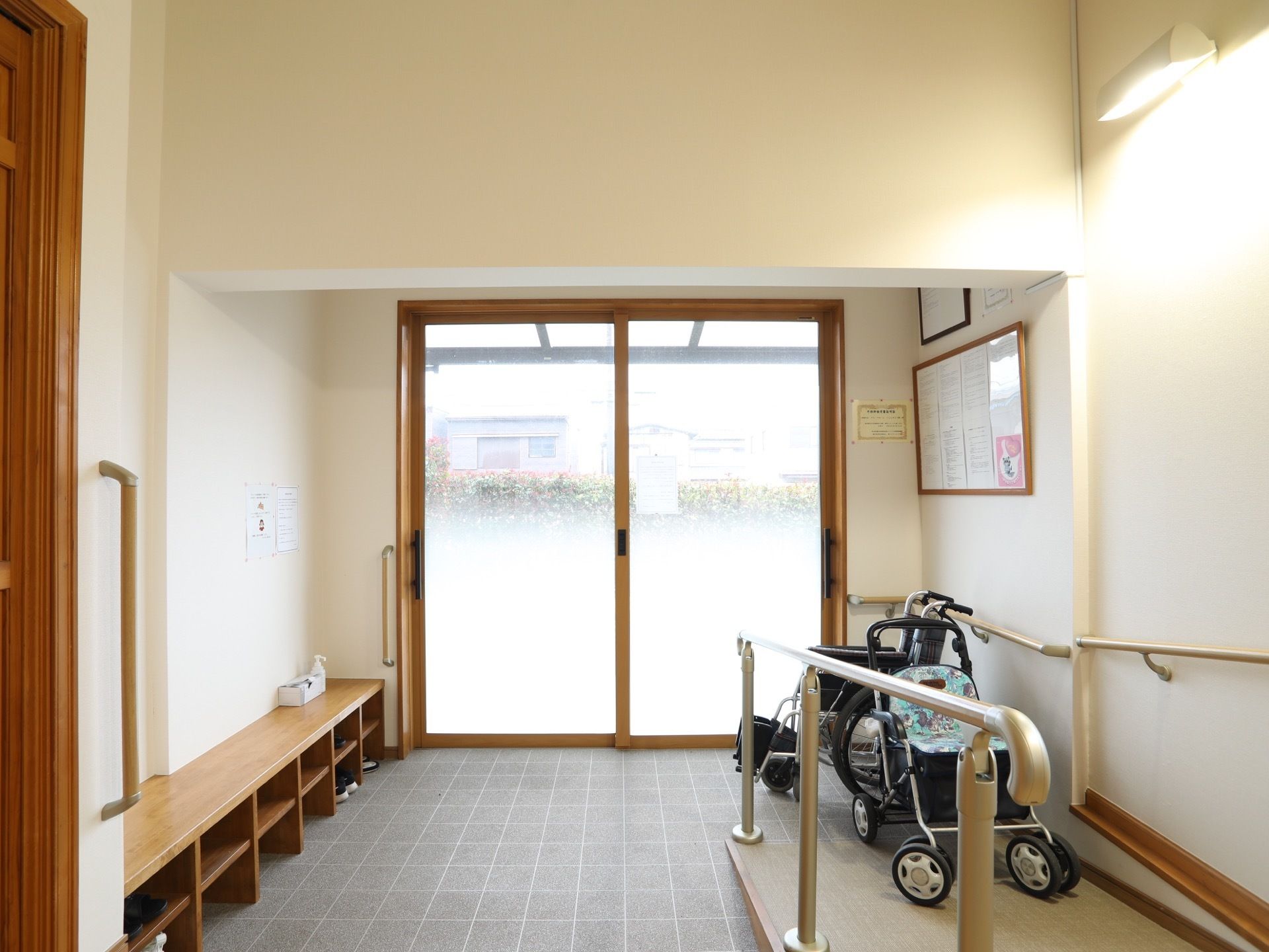 スロープ付きの玄関である。床はグレーのタイルが張られている。壁側には、木製の椅子兼靴箱が設置されている。
