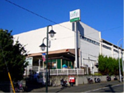施設の近場にあるスーパーには、緑色と白色の看板が建てられている。塀の前には、バイクがたくさん並んでいる。