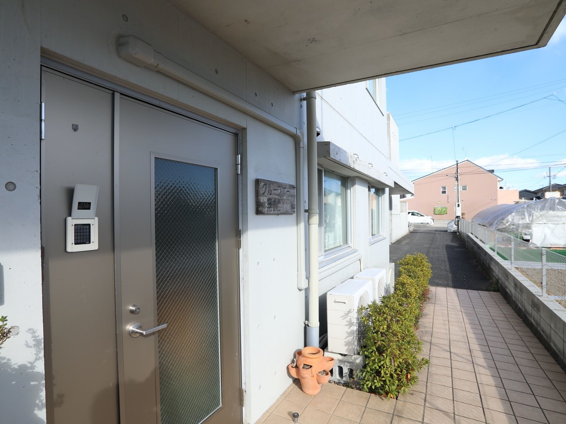 玄関前の通路は、幅が広くすっきりとした空間となっている。隣の敷地の畑との間には、背の低いフェンスが設けられている。