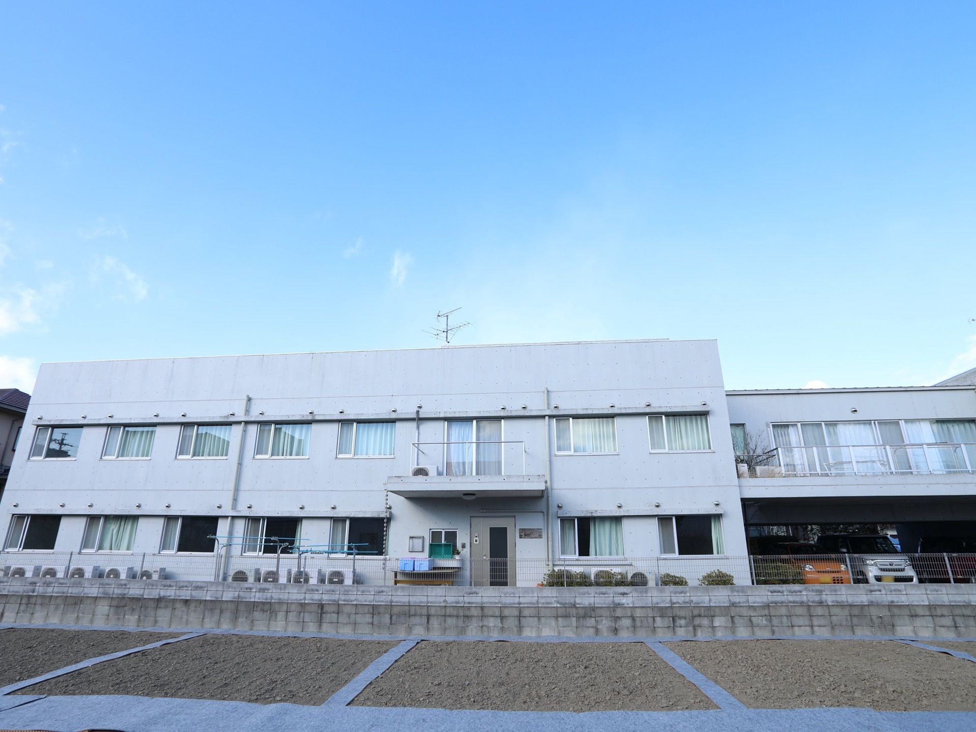 ライトグレーの２階建て施設の外観である。窓からは畑などの広々とした風景が広がっている。敷地内に洗濯物を干す広いスペースが設けられている。