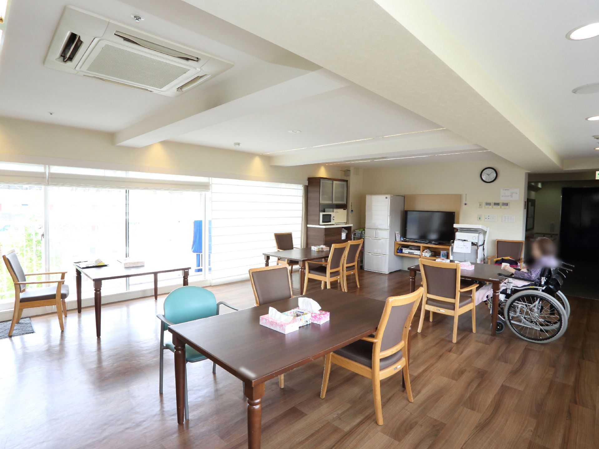 大きな窓がある食堂には冷蔵庫やテレビが並んで置かれている。茶色い机が置かれている周りには椅子が並べられている。