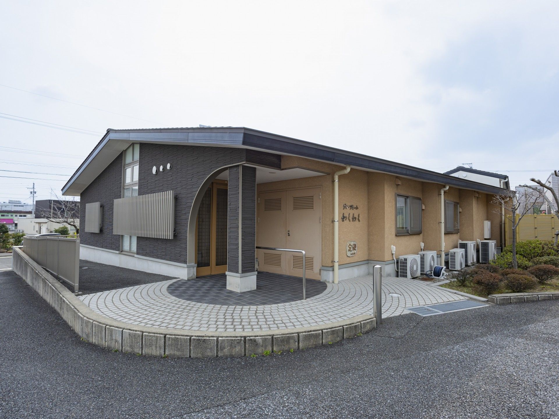 石でできた塀の上に建っている平屋建ての建物はオレンジとブラウン系の外壁で敷地内には草木が植えられている。